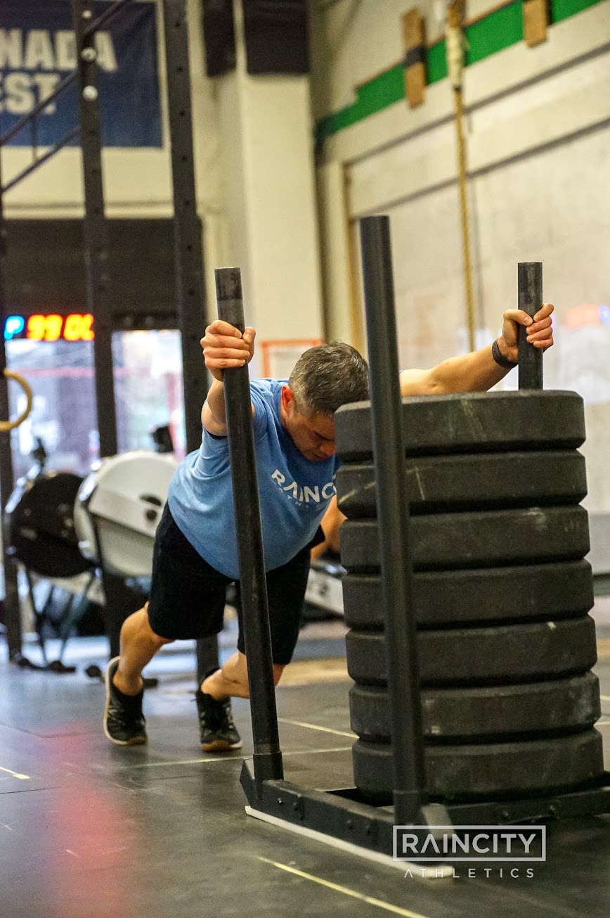 An athlete driving a heavily loaded sled push during a Half Sim heat, race clock running behind them